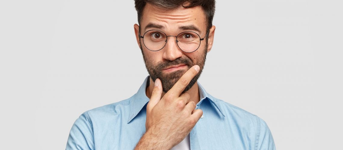 image-hesitant-unshaven-european-man-with-thick-beard-holds-chin-purses-lips-with-clueless-expressions_273609-17361